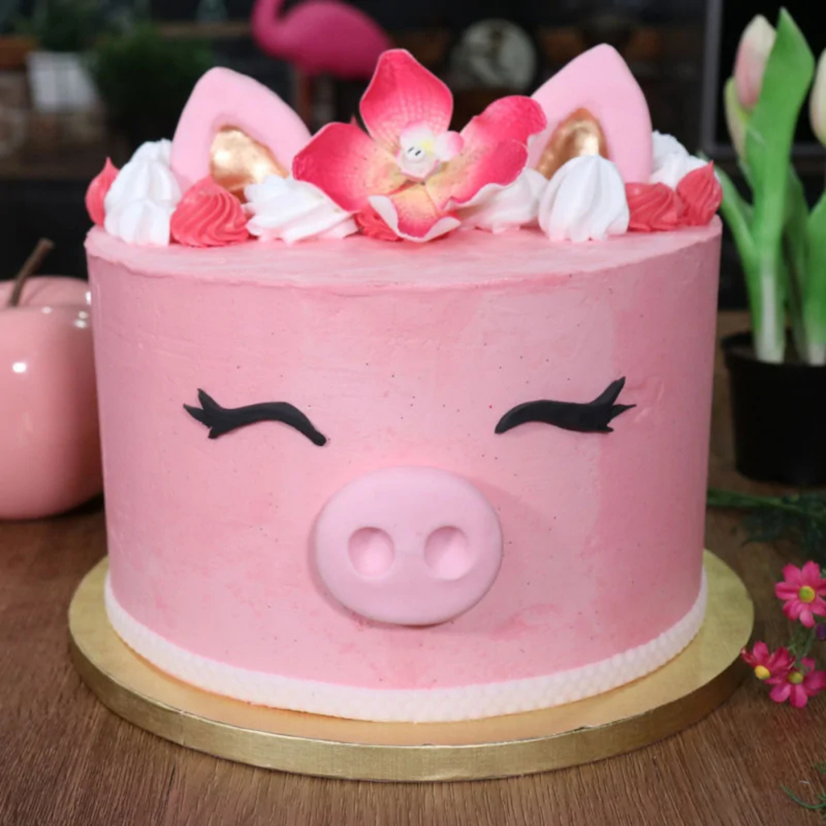 Gâteau rose décoré en forme de cochon avec fleurs, oreilles et détails en pâte à sucre, présenté sur un plateau rond doré