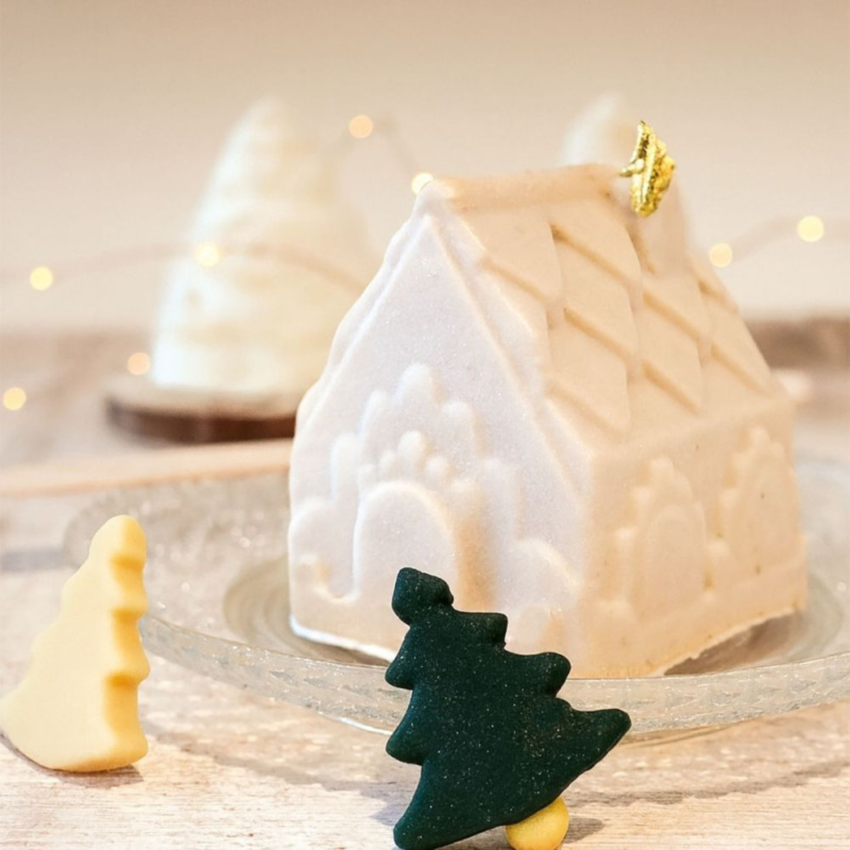 Entremets individuel en forme de chalet de Noël blanc avec détails architecturaux en relief et décoration dorée, accompagné de petits sapins décoratifs.