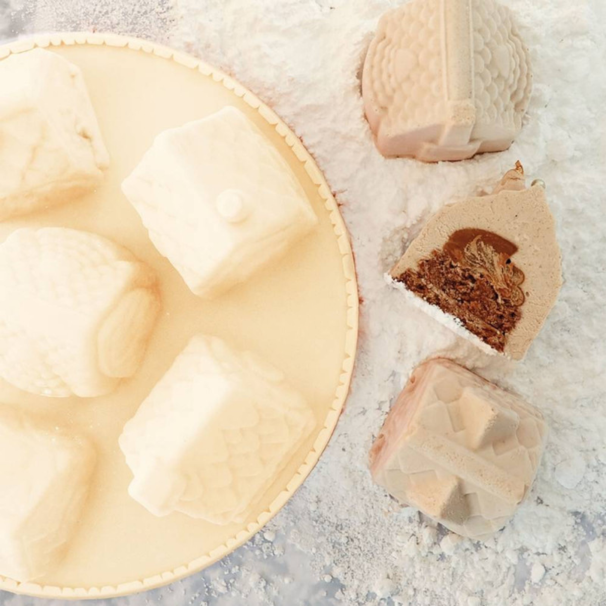 Assortiment d'entremets blancs en forme de chalets de Noël 3D, présentés sur un plateau avec une coupe dévoilant leur garniture au chocolat.