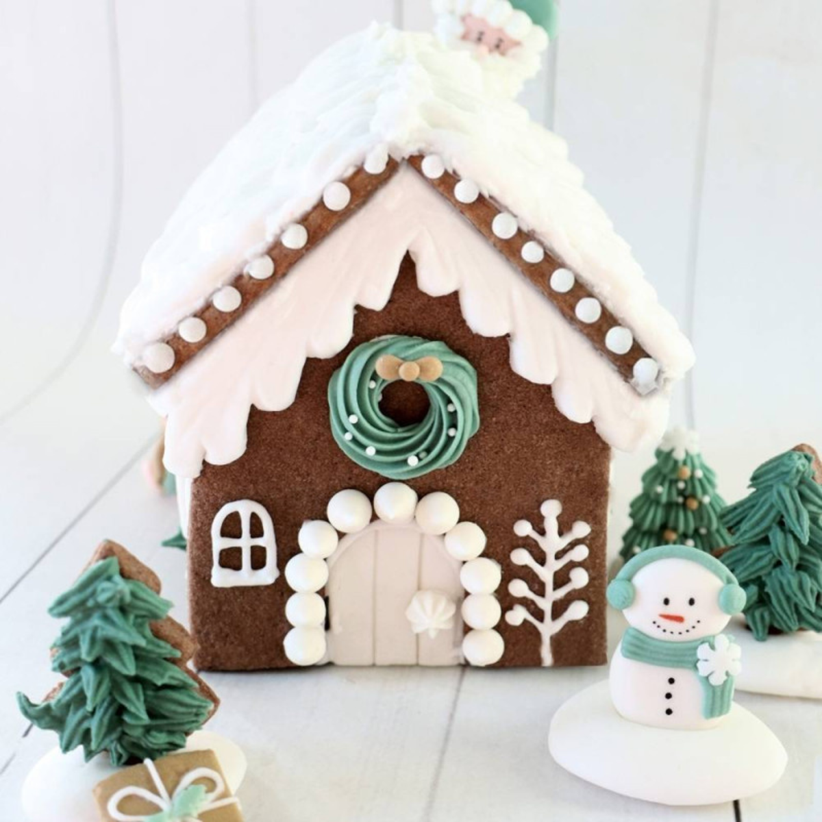 Maison en pain d’épices de Noël décorée de glaçage blanc, couronne verte, sapins et bonhomme de neige, réalisée avec le kit ScrapCooking.