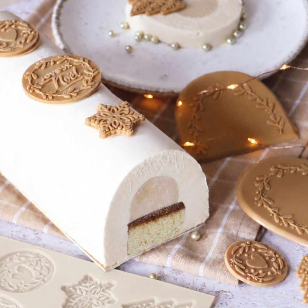 Bûche de Noël blanche en forme de demi-cylindre, décorée de motifs de Noël dorés, avec une coupe laissant apparaître un insert et un biscuit.