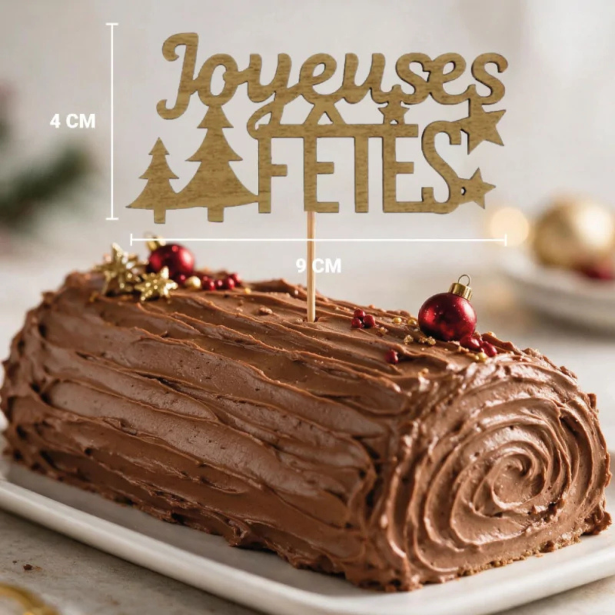 Décor à piquer Joyeuses Fêtes doré de 9 × 4 cm, présenté sur une bûche de Noël au chocolat avec boules rouges et étoiles dorées.