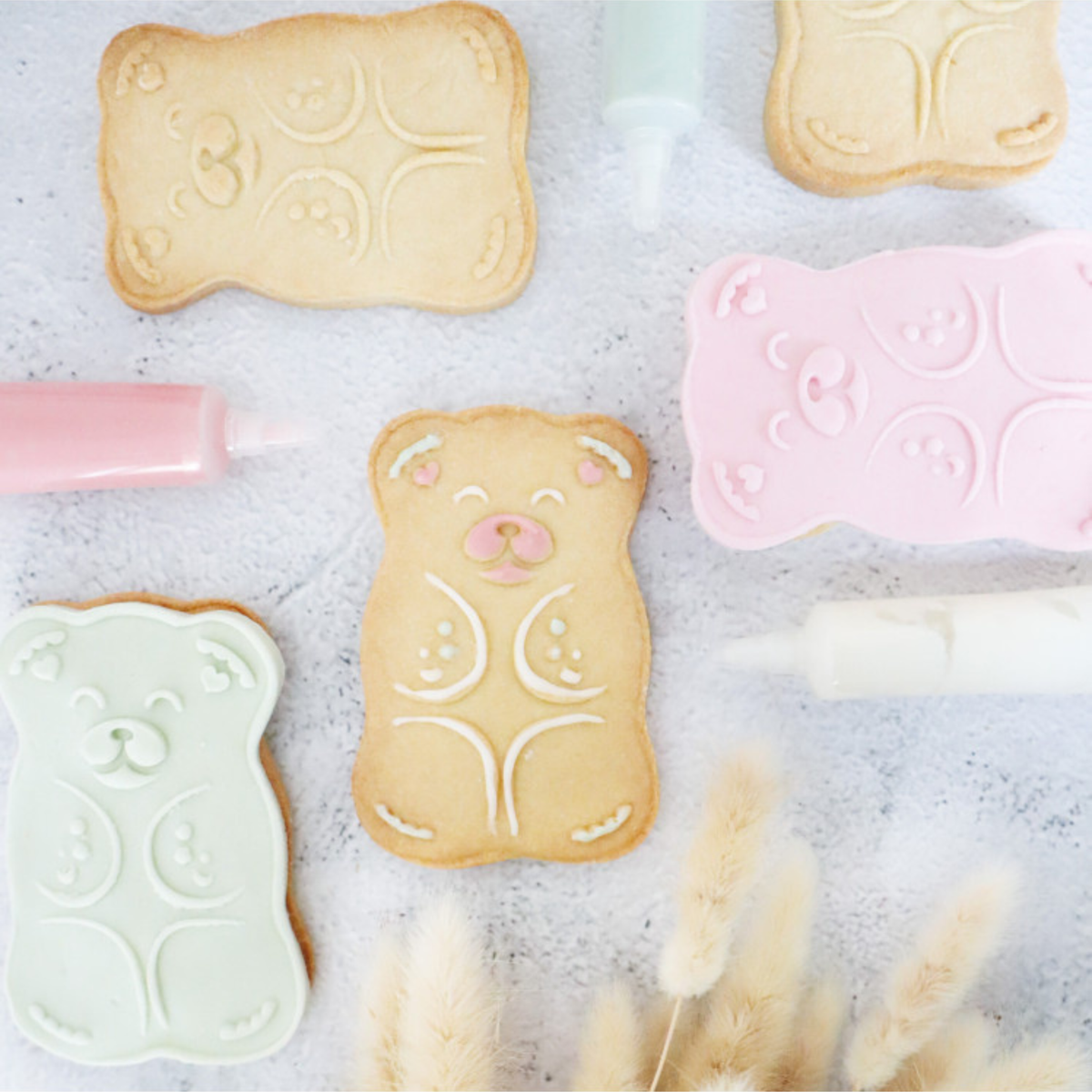 Biscuits sablés en forme d'ourson décorés de glaçage blanc et rose, accompagnés d'embosseurs pastel et de stylos de décoration pâtissière.