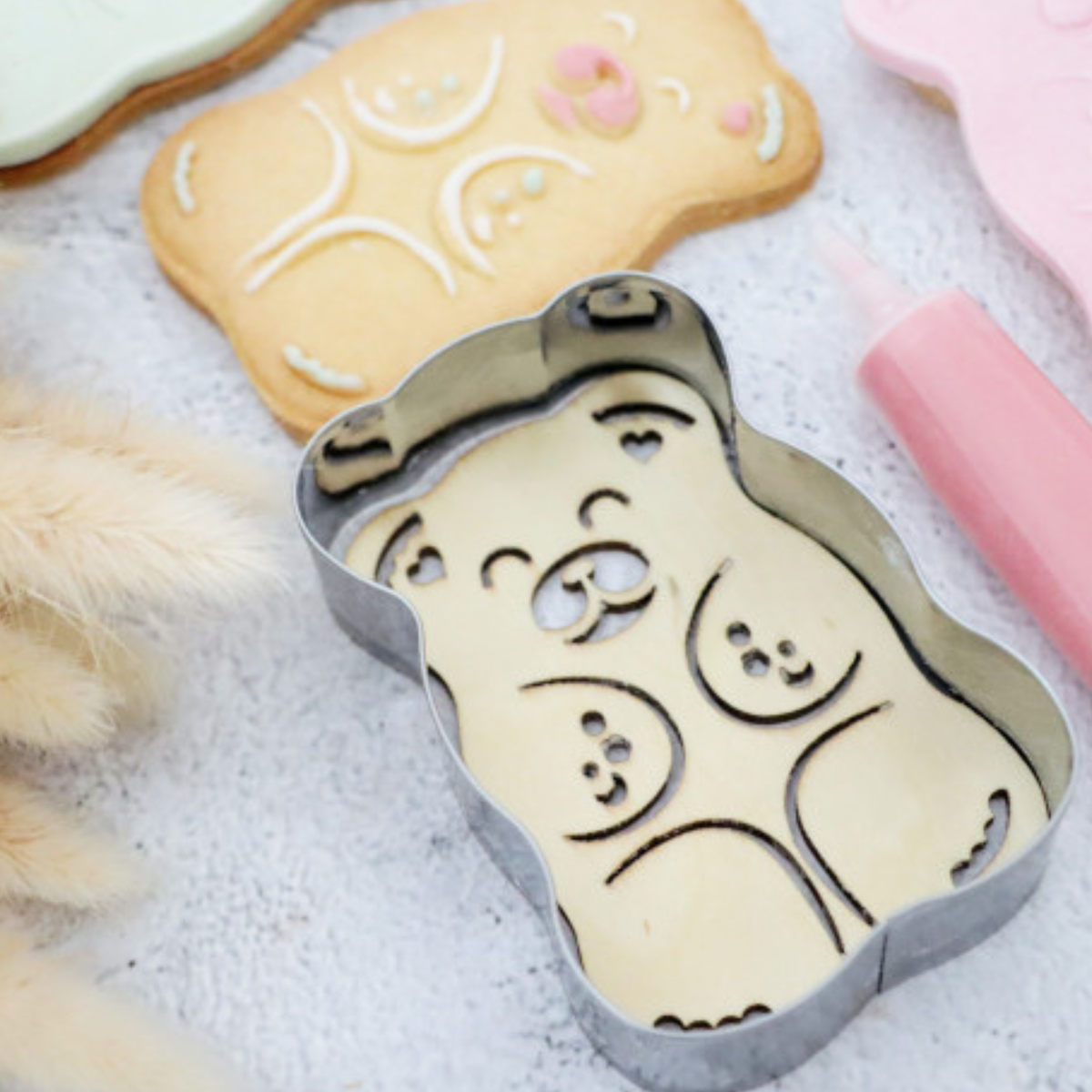 Découpoir embosseur ourson en inox posé sur une pâte à biscuit, entouré de biscuits décorés, de glaçages pastel et d'un stylo pâtissier rose.