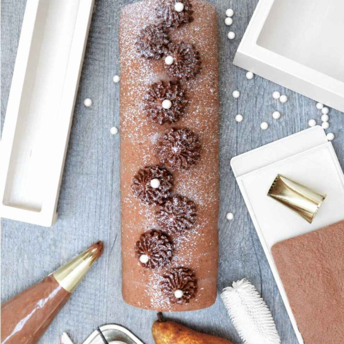 Bûche de Noël au chocolat décorée de rosaces de crème chocolatée, de perles blanches et de sucre glace, accompagnée d'un tapis en silicone blanc ScrapCooking et d'accessoires de pâtisserie.